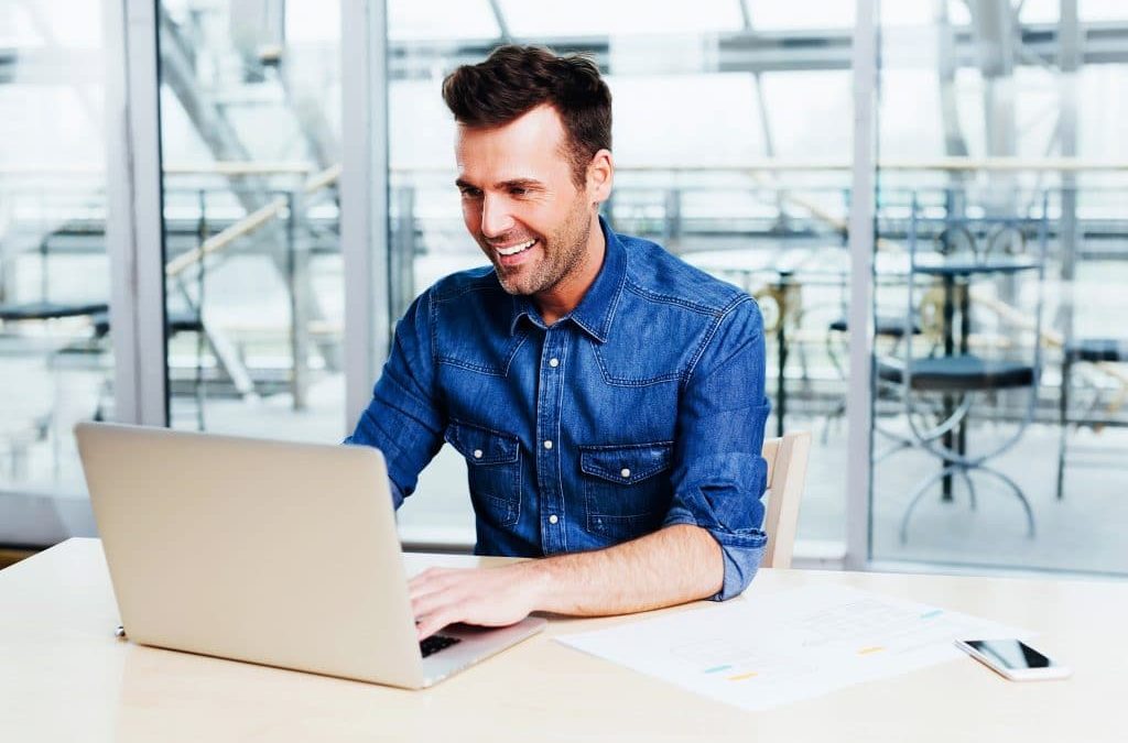 Middle aged man, in denim short, smiling while typing on his laptop