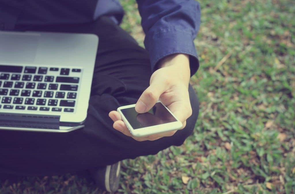 Man siting on grass, with laptop on his lap and a mobile phone in his hand